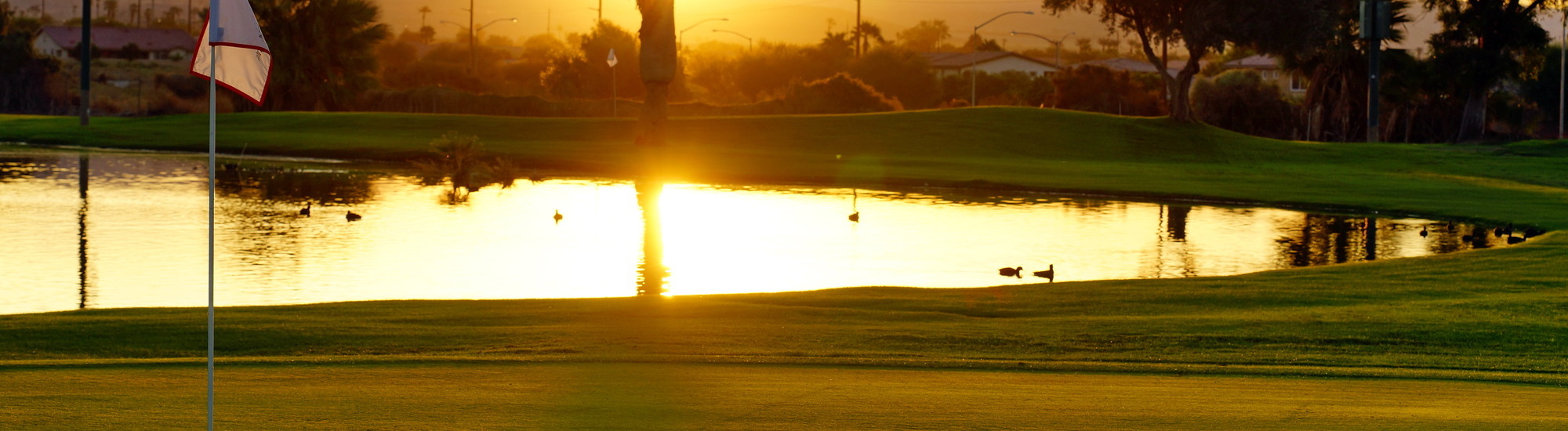 Sunset over a golf course with a pond and ducks, featuring a flag in the foreground.
