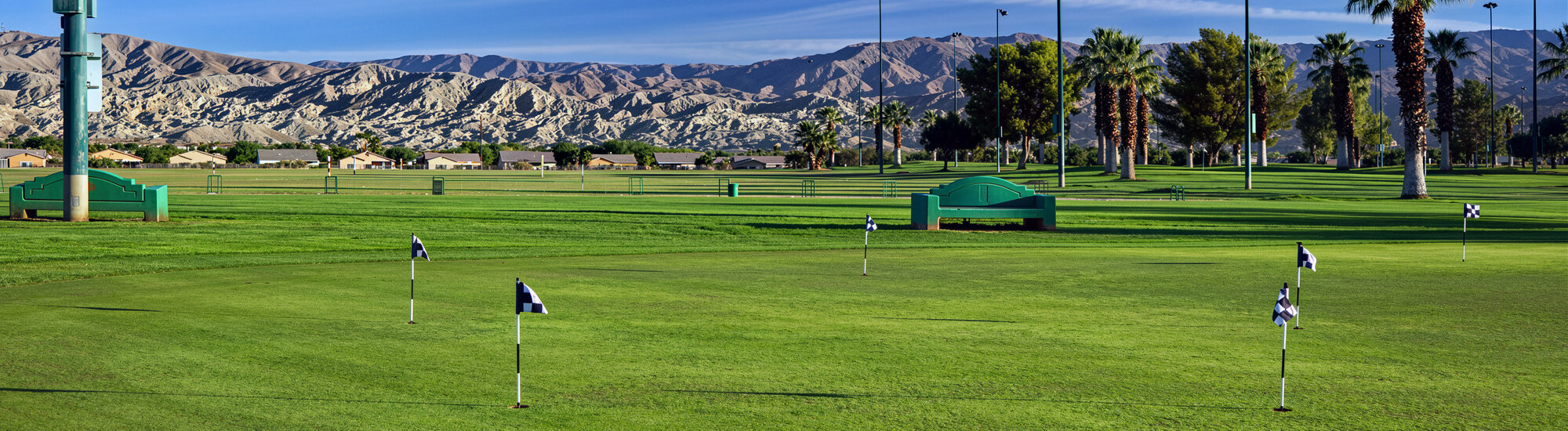 A golf course with multiple flags on the green, surrounded by palm trees and mountains in the background.