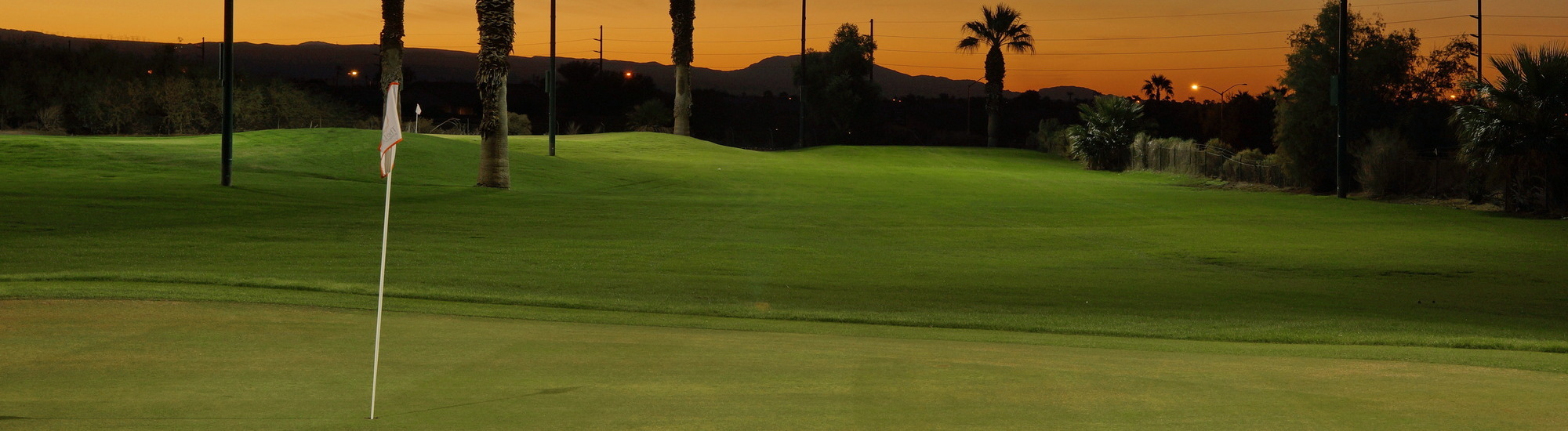 Golf course at sunset with a flag in the foreground and palm trees in the background.
